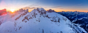 Aerial winter view of Passo Giau at dawn, with the sun rising behind Forcella Giau and Monte Cernera visible on the right. Snow-covered alpine slopes glow in the early morning light. Dolomites, Colle Santa Lucia municipality, Belluno province, Veneto region, Italy