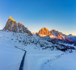 Aerial winter view of Passo Giau at sunrise, with the snow-covered alpine road winding below Ra Gusela and the Tofane group glowing in the first light. Dolomites, Colle Santa Lucia municipality, Belluno province, Veneto region, Italy