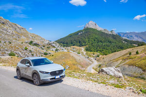 Crossover car driving along the road crossing Mount Visocica plateau, with the rocky ridges of Mount Puzim in the background, Herzegovina-Neretva Canton, Bosnia and Herzegovina Crossover car driving along the road crossing Mount Visocica plateau, with the rocky ridges of Mount Puzim in the background, Herzegovina-Neretva Canton, Bosnia and Herzegovina
