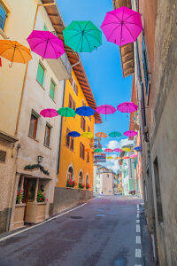 Colorful umbrellas suspended above a narrow cobblestone street lined with bright facades and flowered windows in Castello Tesino. Autonomous Province of Trento, Trentino, Italy Colorful umbrellas suspended above a narrow cobblestone street lined with bright facades and flowered windows in Castello Tesino. Autonomous Province of Trento, Trentino, Italy