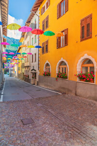 Colorful umbrellas suspended above a narrow cobblestone street lined with bright facades and flowered windows in Castello Tesino. Autonomous Province of Trento, Trentino, Italy Colorful umbrellas suspended above a narrow cobblestone street lined with bright facades and flowered windows in Castello Tesino. Autonomous Province of Trento, Trentino, Italy