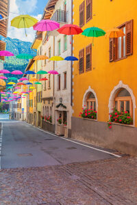 Colorful umbrellas suspended above a narrow cobblestone street lined with bright facades and flowered windows in Castello Tesino. Autonomous Province of Trento, Trentino, Italy Colorful umbrellas suspended above a narrow cobblestone street lined with bright facades and flowered windows in Castello Tesino. Autonomous Province of Trento, Trentino, Italy