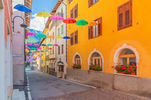 Colorful umbrellas suspended above a narrow cobblestone street lined with bright facades and flowered windows in Castello Tesino. Autonomous Province of Trento, Trentino, Italy Colorful umbrellas suspended above a narrow cobblestone street lined with bright facades and flowered windows in Castello Tesino. Autonomous Province of Trento, Trentino, Italy