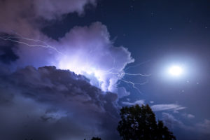 A striking storm cloud from which a lightning bolt emerges with a tree in the foreground and the moon in the clear sky next to it on the island of Sardinia, Italy A striking storm cloud from which a lightning bolt emerges with a tree in the foreground and the moon in the clear sky next to it on the island of Sardinia, Italy