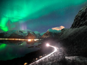 Northern lights over Bergsbotn on Senja island in Norway at night. Meanwhile, a car drives along a road with snow-covered peaks in the background Northern lights over Bergsbotn on Senja island in Norway at night. Meanwhile, a car drives along a road with snow-covered peaks in the background