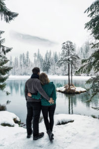 A couple is in Switzerland on Lake Cresta in winter. In the background you can see the mountains, a small island with fir trees and lots of snow. A couple is in Switzerland on Lake Cresta in winter. In the background you can see the mountains, a small island with fir trees and lots of snow.