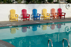 Colorful Adirondack chairs lined up beside a calm swimming pool reflecting the chairs and clear blue sky.