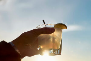 Hand holding a cocktail glass against a sunny sky backdrop with a slice of lemon on the rim.