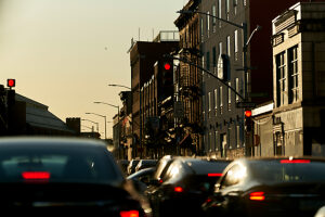 Evening traffic on a city street with red stoplights and the sun setting behind buildings casting a warm glow.