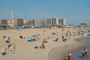 Sandy beach bustling with people on a sunny day, with an amusement park and city skyline in the background.