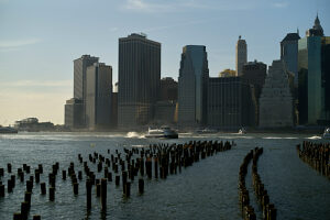Sunset view of the New York City skyline with silhouetted pier remains in the foreground and boats on the water.