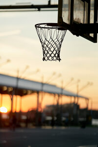 Sunset view through a basketball hoop at an outdoor court with a blurred cityscape in the background.