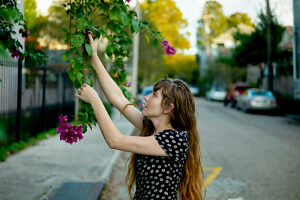 A woman in a black dress with white flowers reaches out to touch purple flowers on a sunny suburban street.