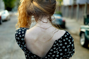 A woman with curly red hair wearing a black dress with white flowers stands with her back to the camera on a city street.