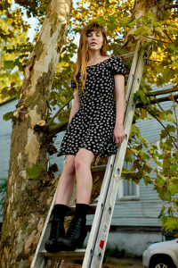 A woman in a black dress with white flowers on it, stands on a ladder leaning against a tree, with a serene expression.
