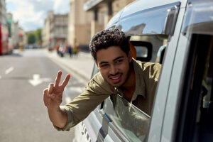 Smiling young man giving a peace sign from the driver's side of a silver car on a sunny city street.