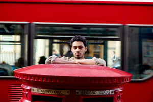 Man leaning on a red Royal Mail postbox with a blurred double-decker bus in the background.