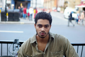 A young man with curly hair looks calmly at the camera, with a blurred city street scene in the background.