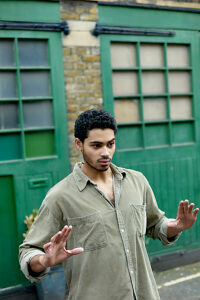 A young man with curly hair stands in front of a green garage door, gesturing with his hands and looking away from the camera.