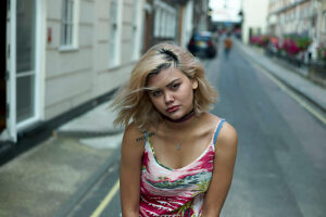 A young woman with blond highlights and a tattoo on her arm stands in a narrow city street, giving a thoughtful look.
