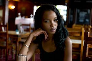 Young woman posing thoughtfully at a wooden table in a dimly lit bar with a glass nearby.