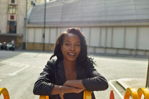 Smiling woman leaning on a yellow barrier on a sunny city street.