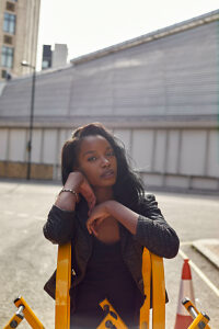 Confident young woman stands with her hand on her cheek beside a yellow metal barrier in an urban setting with buildings and a traffic cone in the background.