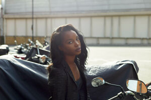 A young woman with long hair is sitting on a motorcycle, looking over her shoulder with a focused expression, in an urban parking area during daylight.