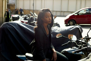 Woman sitting on a motorcycle parked on a city street, with cars and pedestrians in the background.