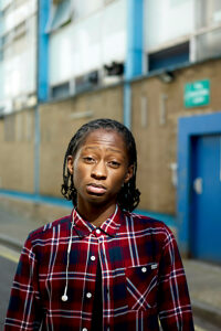 Young adult with dreadlocks wearing a red plaid shirt standing on an urban street with buildings in the background
