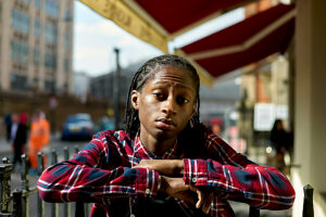 Confident young adult leaning on a railing under a red awning with an urban street in the background.