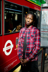 A young adult stands confidently in front of a London bus at night, dressed in a casual plaid shirt with a focused expression.