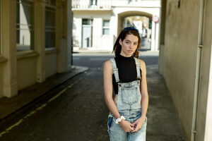 Young woman in denim overalls standing in a sunlit alleyway, looking thoughtful.