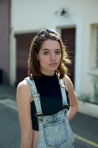 Young woman with hairband on her head wearing denim overalls and a black top standing in front of a garage door.