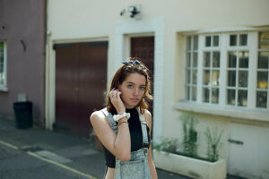 Young woman standing by a white doorway in an alley, with a thoughtful expression as she touches her hair.