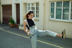 Young woman in denim overalls playfully kicking in a sunlit urban street.