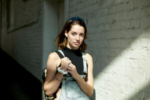 Young woman with a denim overall posing against a white brick wall.