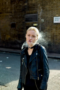 Woman exhaling smoke while standing against a brick wall in sunlight wearing a black leather jacket.