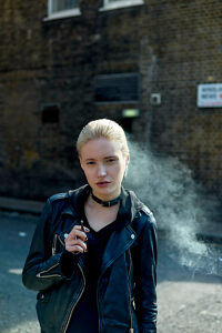A blonde woman exhales smoke while holding a cigarette, standing in front of a brick wall with a 'No Parking' sign.