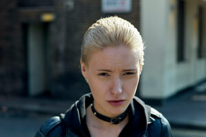 Close-up of a young woman with short blonde hair wearing a black leather jacket and a choker on a sunny street.