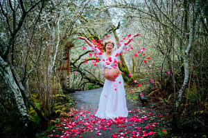 Pregnant woman in white dress celebrating in forest surrounded by pink petals. 