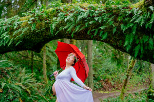 Pregnant woman in white dress with red umbrella under a lush forest archway. 