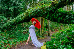 Pregnant woman in white dress with red umbrella under a lush forest archway. 