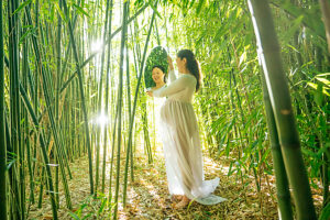 Pregnant woman in white dress holds a mirror in a serene bamboo forest. 