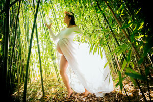 Pregnant woman in long dress stands amid lush green bamboo forest, holding a glowing orb. 
