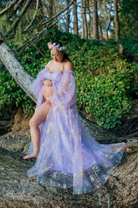 Pregnant woman in floral gown sits on a tree with lush greenery in the background. 