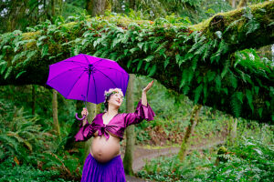 Pregnant woman in purple with umbrella smiles under lush green forest canopy. 