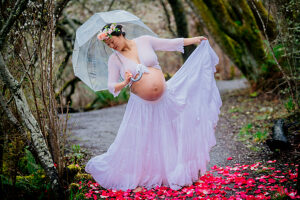 Pregnant woman in flowy white dress holds umbrella on forest path with pink flower petals. 