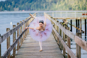 Ballerina performing a graceful pose on a wooden dock with scenic water and forest backdrop. WA, USA