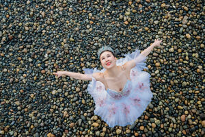 Ballerina in a tutu joyfully poses with arms outstretched on a pebble beach. WA, USA